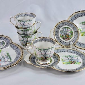 Puede incluir: Un juego de t&eacute; vintage con tazas, platillos y platos de servir. El juego presenta un dise&ntilde;o de paisaje con &aacute;rboles y flores, enmarcado por un borde gris y dorado. Las tazas est&aacute;n apiladas y los platos est&aacute;n dispuestos.