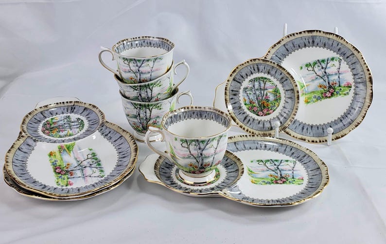 Puede incluir: Un juego de t&eacute; vintage con tazas, platillos y platos de servir. El juego presenta un dise&ntilde;o de paisaje con &aacute;rboles y flores, enmarcado por un borde gris y dorado. Las tazas est&aacute;n apiladas y los platos est&aacute;n dispuestos.