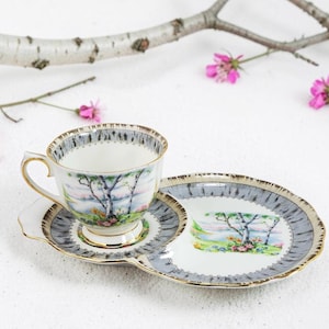 Puede incluir: Un juego de taza y platillo de t&eacute; vintage con un dise&ntilde;o de paisaje. La taza, el platillo y el plato que lo acompa&ntilde;a presentan un borde gris y dorado. El dise&ntilde;o del paisaje incluye &aacute;rboles y una masa de agua. Flores rosas y una rama en el fondo.