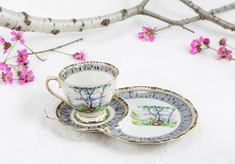 Puede incluir: Un juego de taza y platillo de t&eacute; vintage con un dise&ntilde;o de paisaje. La taza, el platillo y el plato que lo acompa&ntilde;a presentan un borde gris y dorado. El dise&ntilde;o del paisaje incluye &aacute;rboles y una masa de agua. Flores rosas y una rama en el fondo.