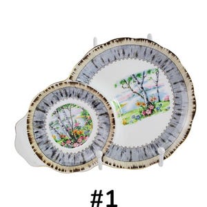 Puede incluir: Dos platos de servir vintage con un dise&ntilde;o de paisaje. Los platos tienen una base blanca con una escena pintada de &aacute;rboles, flores y una masa de agua. Los bordes son grises con un borde marr&oacute;n. El texto "#1" est&aacute; debajo.