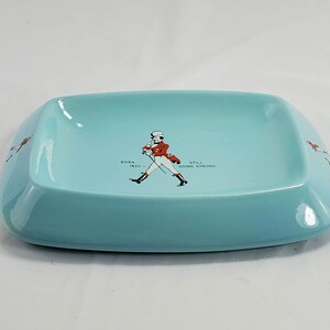 May include: A rectangular, light blue ashtray with rounded corners. The center features a graphic of a man in a red coat and top hat, with the text "BORN 1820 - STILL GOING STRONG". The sides have a graphic of a running man.