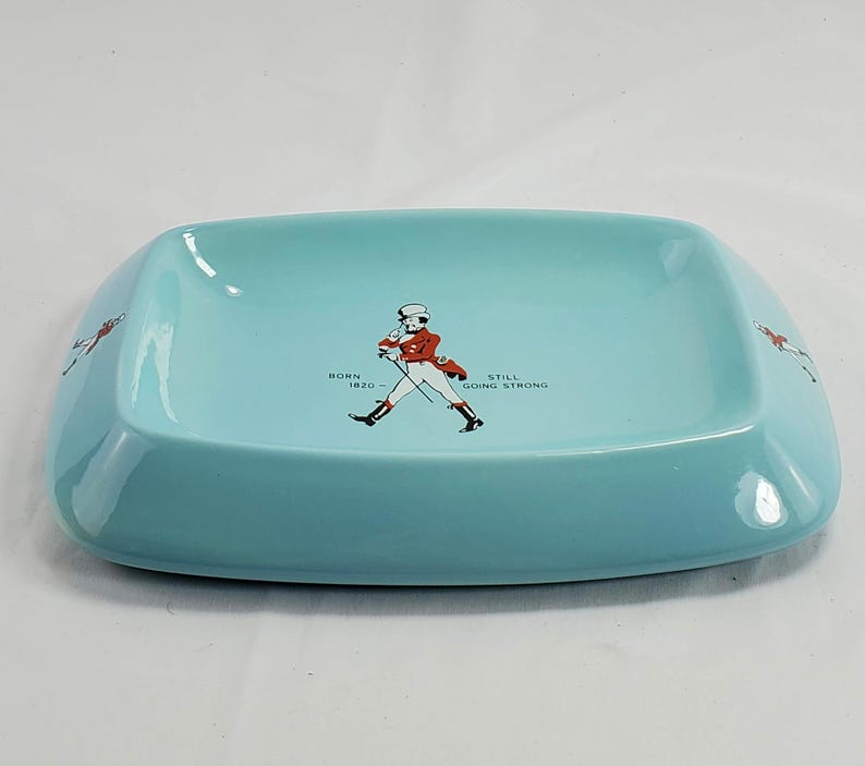 May include: A rectangular, light blue ashtray with rounded corners. The center features a graphic of a man in a red coat and top hat, with the text "BORN 1820 - STILL GOING STRONG". The sides have a graphic of a running man.