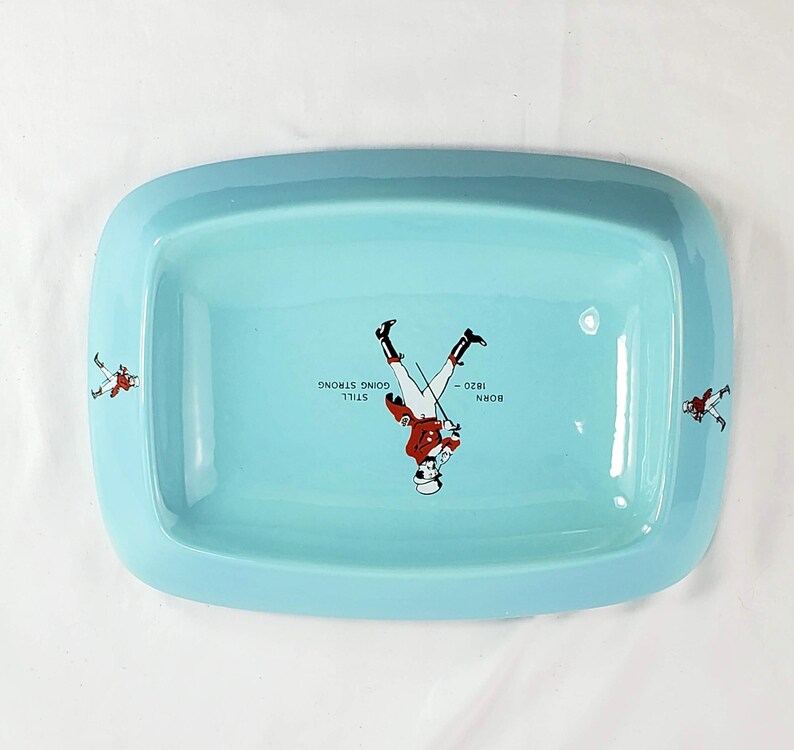 May include: A rectangular, light blue ceramic tray with rounded corners. The tray features a central illustration of a soldier and the text "BORN 1820 - STILL GOING STRONG". Additional soldier illustrations are on the tray's edges.