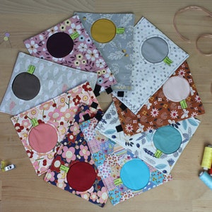 May include: Nine fabric pouches with different patterns and colors. Each pouch has a circular opening with a contrasting fabric circle inside. The pouches are arranged in a circle on a wooden surface.