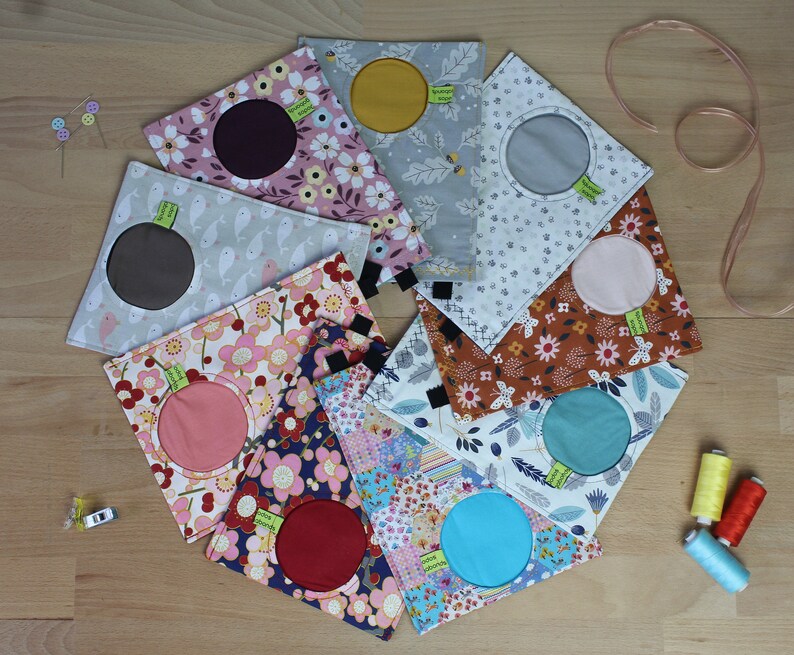 May include: Nine fabric pouches with different patterns and colors. Each pouch has a circular opening with a contrasting fabric circle inside. The pouches are arranged in a circle on a wooden surface.