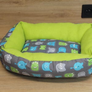 May include: A small pet bed with a gray exterior patterned with blue and green cartoon monsters. The interior and bolster are bright green. The bed is oval-shaped and sits on a wooden surface.
