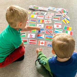 Puede incluir: Dos ni&ntilde;os juegan un juego con un conjunto de banderas coloridas que representan diferentes pa&iacute;ses. Las banderas est&aacute;n dispuestas sobre una alfombra gris. Un ni&ntilde;o lleva una camisa verde y pantalones rojos, mientras que el otro ni&ntilde;o lleva una camisa azul.