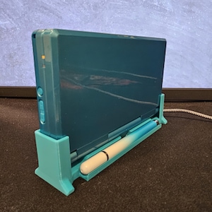 May include: Teal handheld gaming console in a teal stand with a white and blue stylus. The console is open, revealing the screen. The stand is on a dark surface, with a monitor in the background.