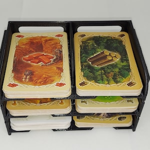 May include: A black plastic card organizer with four tiers for holding game cards. The top tier has two stacks of cards, one with a picture of bricks and the other with a picture of logs.