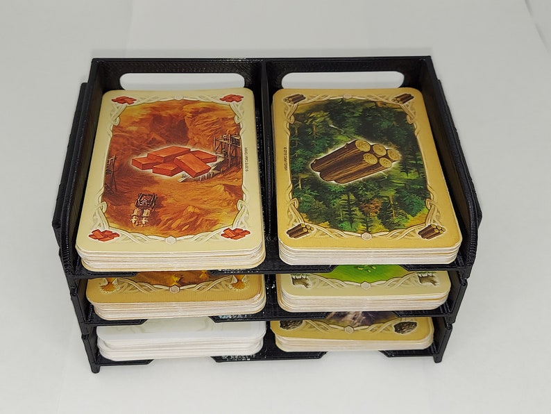 May include: A black plastic card organizer with four tiers for holding game cards. The top tier has two stacks of cards, one with a picture of bricks and the other with a picture of logs.