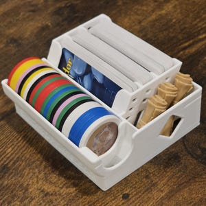 May include: A white plastic game organizer with compartments for game pieces. It holds stacks of cards, colorful tokens, and beige game pieces. The organizer is on a wooden surface.
