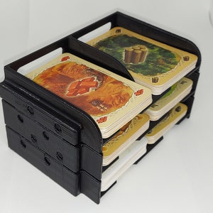 May include: Black plastic card holder with multiple slots for organizing cards. The holder is filled with cards featuring illustrations of different resources, including wood, brick, and sheep.
