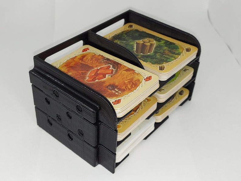 May include: Black plastic card holder with multiple slots for organizing cards. The holder is filled with cards featuring illustrations of different resources, including wood, brick, and sheep.