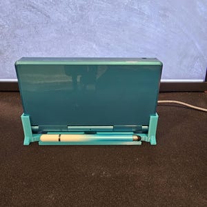 May include: Teal handheld games console with a matching stylus, in a teal stand. The console is closed, showing its back. The stand is designed to hold the console upright, and the stylus is placed in a slot below.