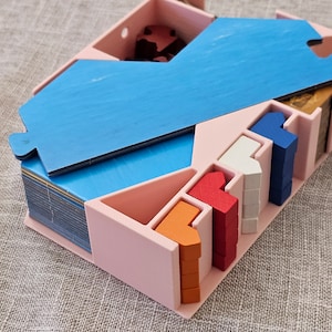 以下が含まれることがあります： 小さなゲームピースを収納するためのピンクのプラスチック製オーガナイザー。コンパートメントには、赤、オレンジ、白、青のウッドピースが入っています。
