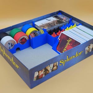 May include: A blue game box with a custom insert for organizing the game pieces. The insert has compartments for cards, tokens, and other game components. The box is labeled "Splendor".