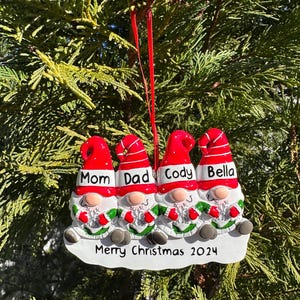 May include: A Christmas ornament featuring four gnomes with red hats and white beards. The gnomes are sitting together and have the names "Mom", "Dad", "Cody", and "Bella" written on their hats. The ornament also has the text "Merry Christmas 2024" written on it.