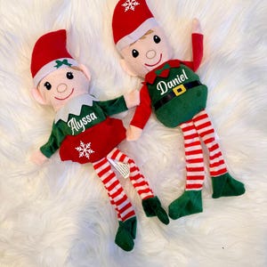 May include: Two plush elf dolls with red and white striped legs, green elf suits, and red hats. The elf on the left has a snowflake on its suit and the name "Alyssa" embroidered on it. The elf on the right has the name "Daniel" embroidered on its suit.