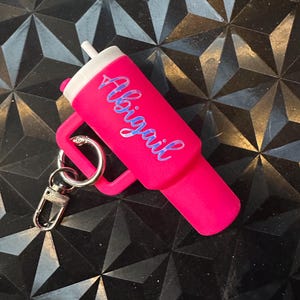 May include: A pink keychain with a small, personalized tumbler. The tumbler has a white lid and a straw. The name "Abigail" is written in blue on the side of the tumbler.