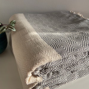 May include: A folded grey and white patterned blanket with a cream border. The blanket has a diamond pattern and is made of a soft, woven material. The blanket is folded neatly, showcasing its texture and design.