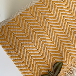 May include: A yellow and white patterned throw blanket with fringe edges. The blanket is made of cotton and has a herringbone pattern.