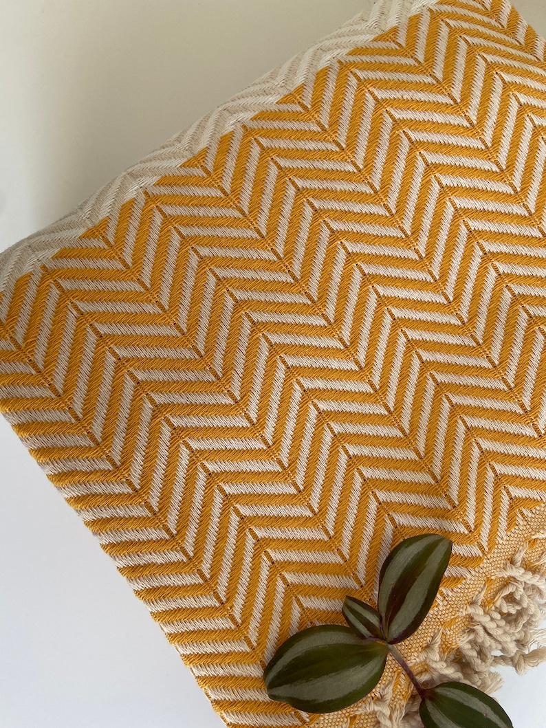 May include: A yellow and white patterned throw blanket with fringe edges. The blanket is made of cotton and has a herringbone pattern.