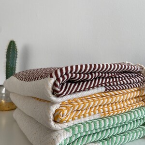 May include: A stack of four woven cotton towels in brown, yellow, green, and white. The towels are folded and stacked on a white surface.