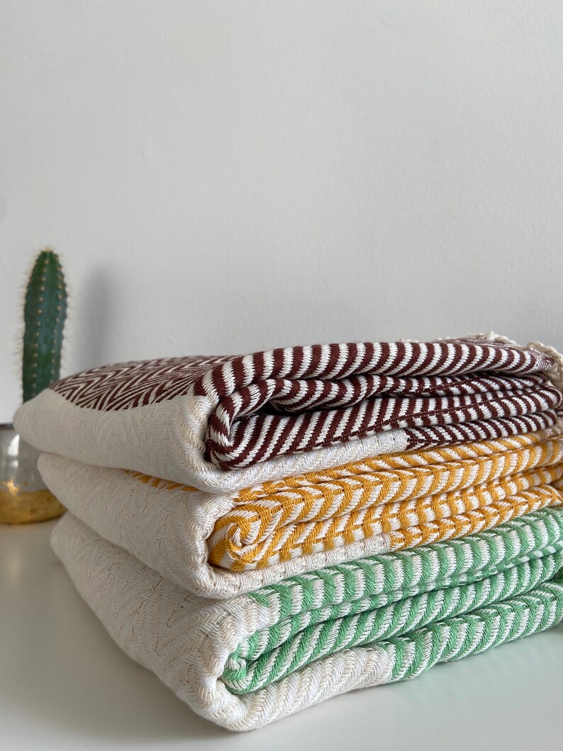May include: A stack of four woven cotton towels in brown, yellow, green, and white. The towels are folded and stacked on a white surface.