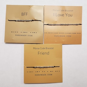 May include: Three Morse code bracelets on tan cards. One says "BFF", another "I Love You", and the third "Friend". Each bracelet has silver beads on a black cord. The cards also say "HANDMADE ITEM".