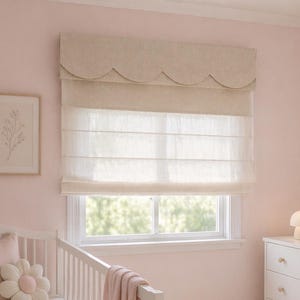May include: A nursery scene with a white crib, a flower-shaped pillow, and a pink blanket. A Roman shade with a scalloped valance hangs above a window. A white dresser and a framed botanical print complete the soft, inviting decor.