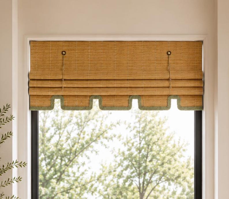 May include: A woven bamboo roman shade featuring a natural tan finish with a scalloped bottom edge trimmed in a muted sage green fabric. The shade includes two vertical cord mechanisms for lifting and is mounted within a window frame.