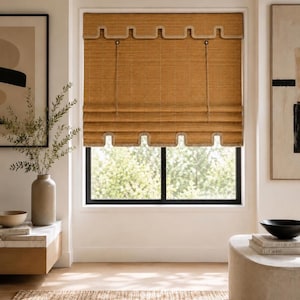 May include: A woven bamboo roman shade in a natural tan color featuring a scalloped edge design along the top valance and the bottom hem. The shade includes thin vertical pull cords and a textured, grid-like pattern across the material.