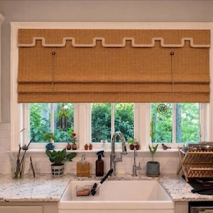 May include: A tan woven roman shade with a decorative cream-colored Greek key trim along the top valance. The shade is mounted inside a white window frame above a kitchen sink, featuring a textured fabric construction with horizontal folds.