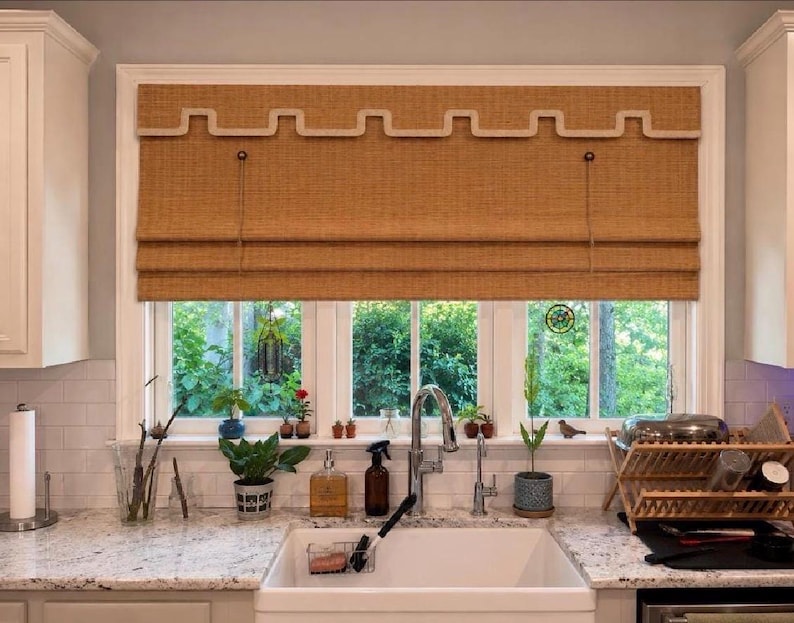 May include: A tan woven roman shade with a decorative cream-colored Greek key trim along the top valance. The shade is mounted inside a white window frame above a kitchen sink, featuring a textured fabric construction with horizontal folds.