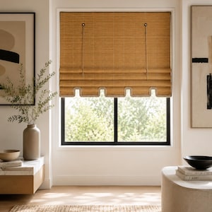 May include: A woven bamboo roman shade with a light brown finish, featuring a scalloped bottom edge with a light-colored trim. The shade is suspended by two thin cords attached to circular hardware at the top, covering a rectangular window frame.