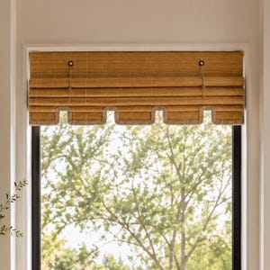 May include: A tan bamboo roll-up window shade featuring a horizontal slatted texture and a scalloped bottom edge trimmed with a light-colored fabric border. The shade is suspended by two thin cords attached to small circular hardware at the top.