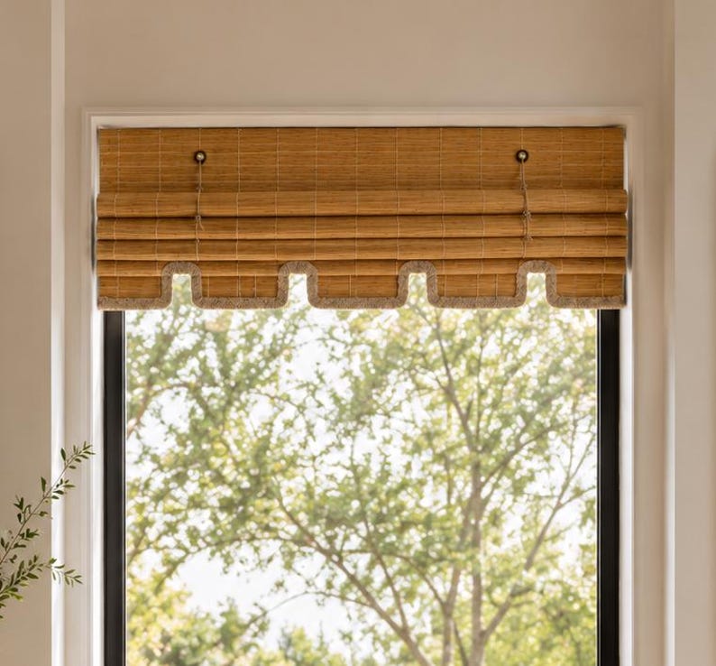 May include: A tan bamboo roll-up window shade featuring a horizontal slatted texture and a scalloped bottom edge trimmed with a light-colored fabric border. The shade is suspended by two thin cords attached to small circular hardware at the top.