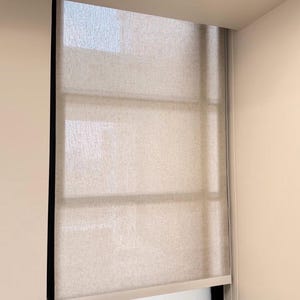 May include: A neutral-toned roller shade is installed in a window. The shade is made of a textured fabric and is partially lowered, revealing a portion of the window. The shade is framed by a black border and a white trim.