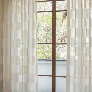 Puede incluir: Cortinas transparentes de color crema con un patrón geométrico en forma de cuadrícula enmarcan una ventana con marco marrón. Las cortinas son semitransparentes, permitiendo el paso de la luz natural. La ventana ofrece vistas a árboles y una pared blanca.
