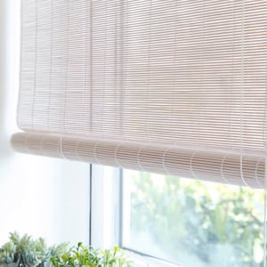 May include: A light beige bamboo window shade, partially rolled up, hangs in front of a window. The shade is made of thin, horizontal bamboo slats held together with vertical string. The bottom edge is rolled up, revealing the window and greenery outside.