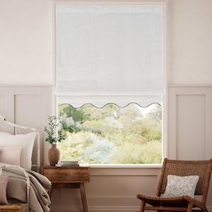 May include: A white Roman shade with a scalloped edge and a dark trim is installed in a window. The window looks out onto a lush green landscape. A wooden nightstand with a vase of flowers and a wicker chair are in the room.