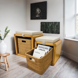May include: Two wooden storage ottomans with beige cushions and woven baskets. The ottomans are in a room with white walls and artwork. One basket is filled with books, including "Modigliani".