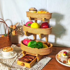 May include: A three-tiered bamboo fruit basket with woven rattan baskets. The top basket holds a small cake and a pastry. The middle basket holds fruit, including apples, pears, and oranges. The bottom basket holds more fruit and a small green plant. The basket is on a wooden table with a white tablecloth.