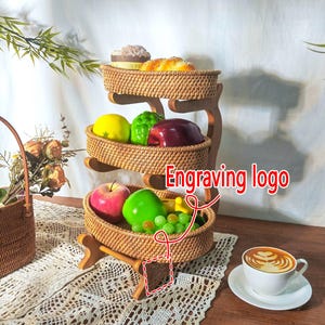 Pode incluir: Um suporte de cesta de frutas de vime tecido de três níveis com uma base de madeira. O suporte está cheio de frutas, incluindo maçãs, uvas e peras. O suporte tem um logotipo gravado no lado.