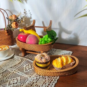 May include: Two woven wicker baskets on a wooden stand. The top basket holds fruit, including a red apple, a yellow banana, a pink pear, and green grapes. The bottom basket holds two pastries. The baskets are on a white lace tablecloth.