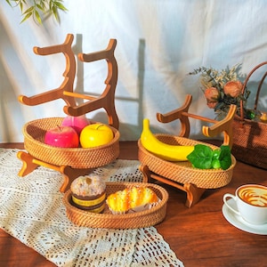 May include: Three woven wicker baskets with wooden stands, two with fruit and one with pastries. The baskets are oval shaped and have a natural brown color. The stands are made of light brown wood and have a simple design.