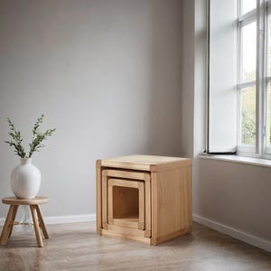 May include: Three wooden nesting tables in a natural wood finish. The tables are square and have a simple design. The largest table is on the bottom, and the smallest table is on top.