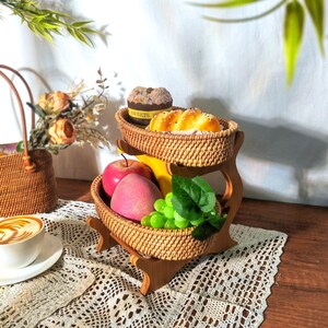 May include: A two-tiered woven wicker fruit and bread basket stand with a wooden base. The top tier holds a loaf of bread and a small cake, while the bottom tier holds apples, grapes, and a banana.