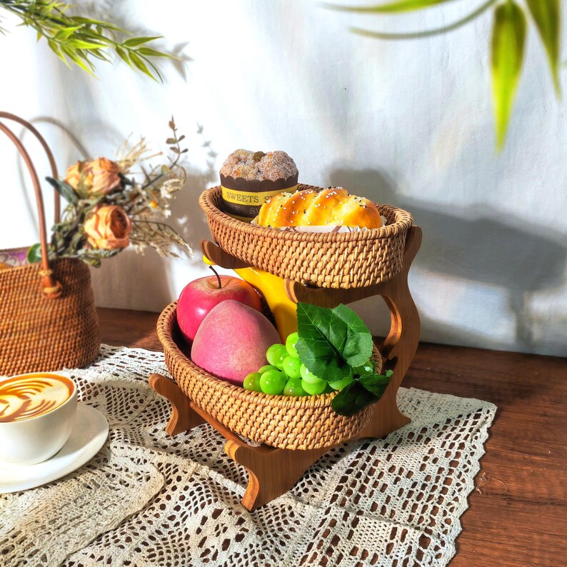 May include: A two-tiered woven wicker fruit and bread basket stand with a wooden base. The top tier holds a loaf of bread and a small cake, while the bottom tier holds apples, grapes, and a banana.